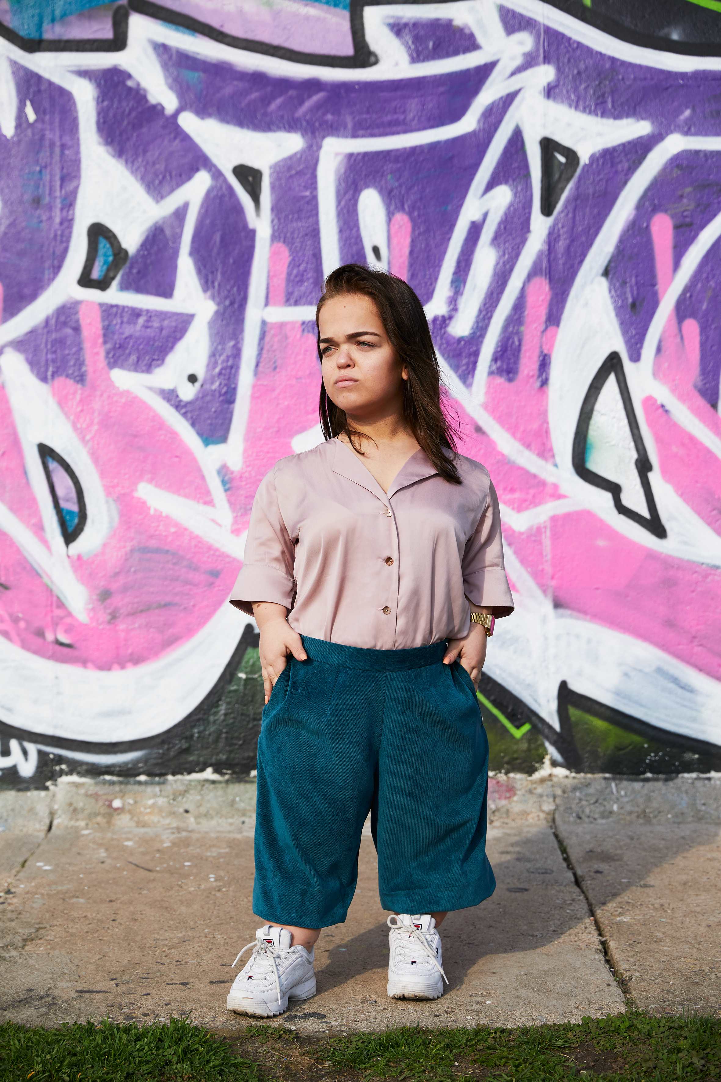 woman with dwarfism in front of grafitti wall wearing matching rose blouse and turquoise pants