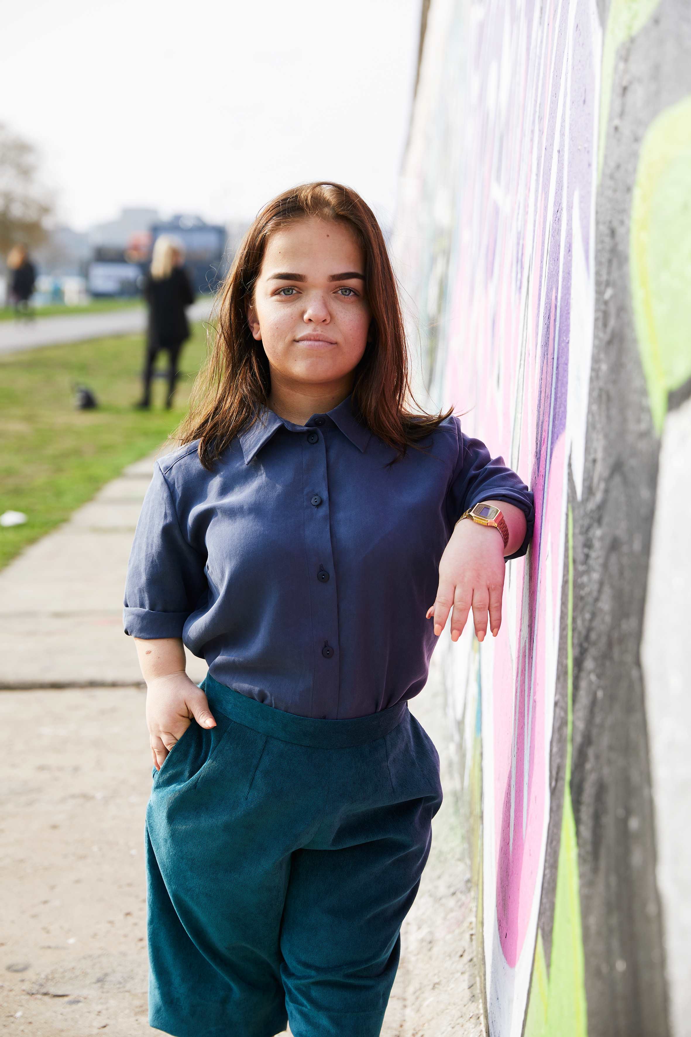 woman with dwarfism leaning casually against grafitti wall, wearing blue blouse