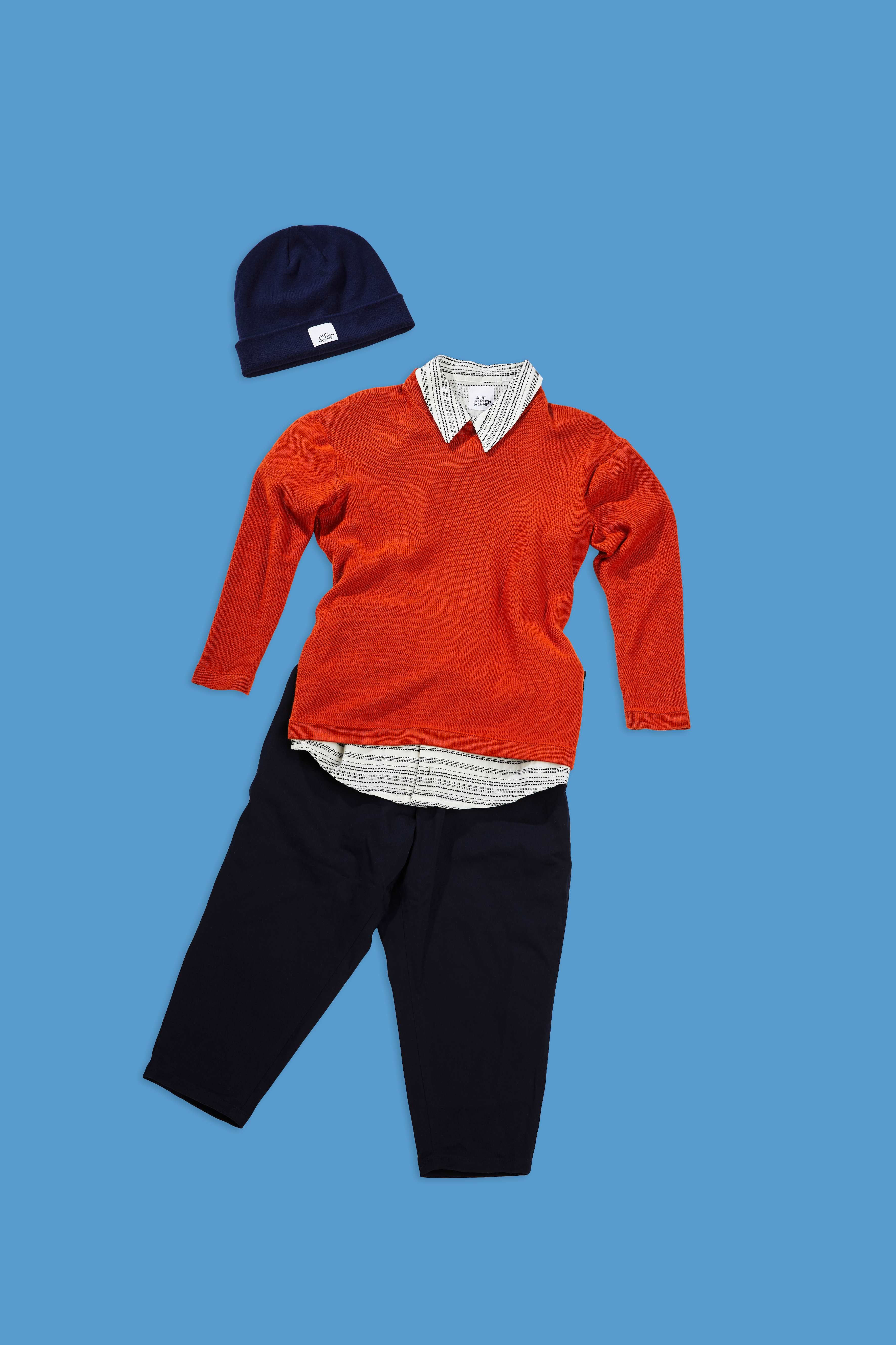 rusty red pullover with striped shirt, black pants and beanie on blue background