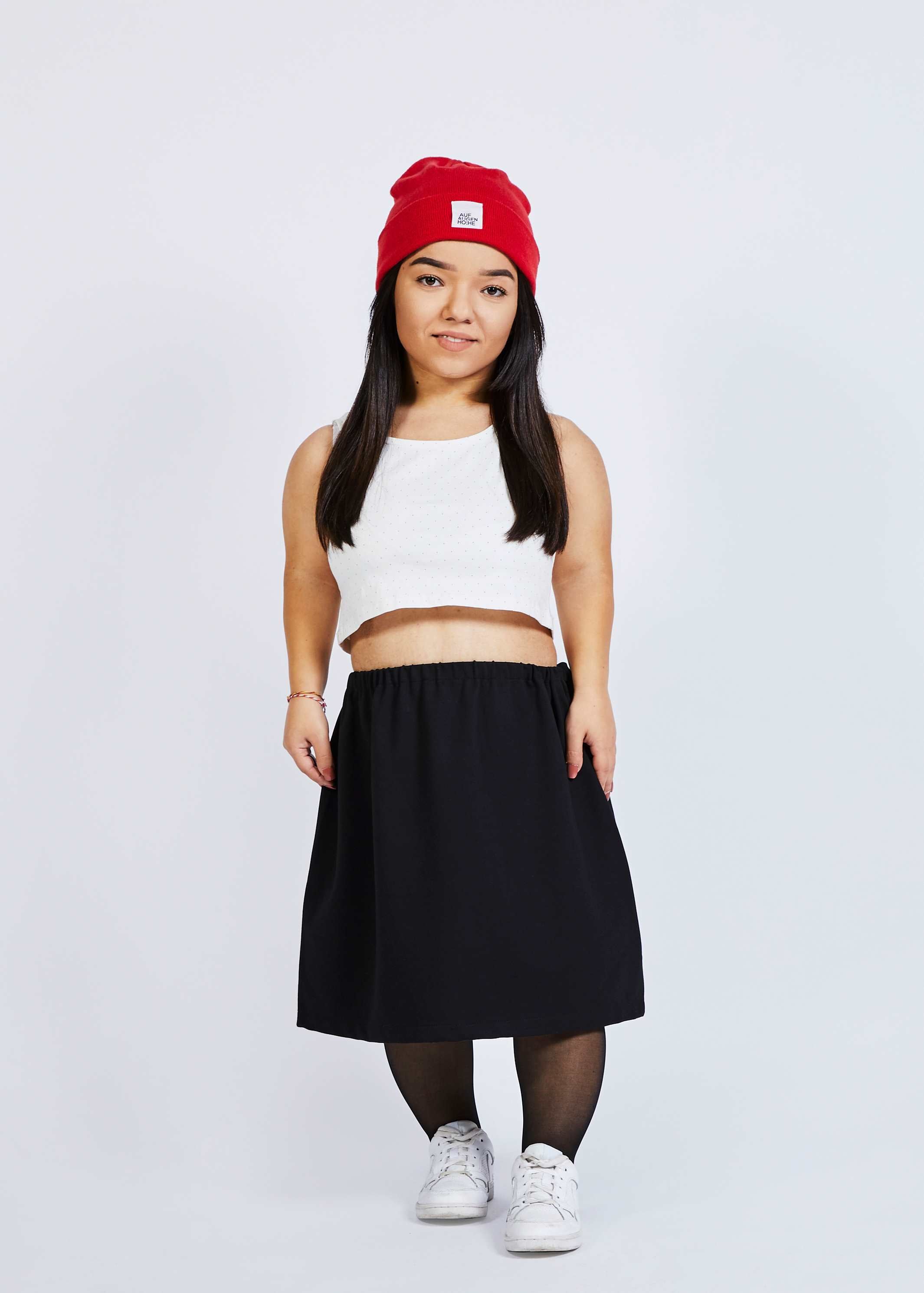 woman with dwarfism wearing black skirt, tights, white croptop, red beanie and white shoes